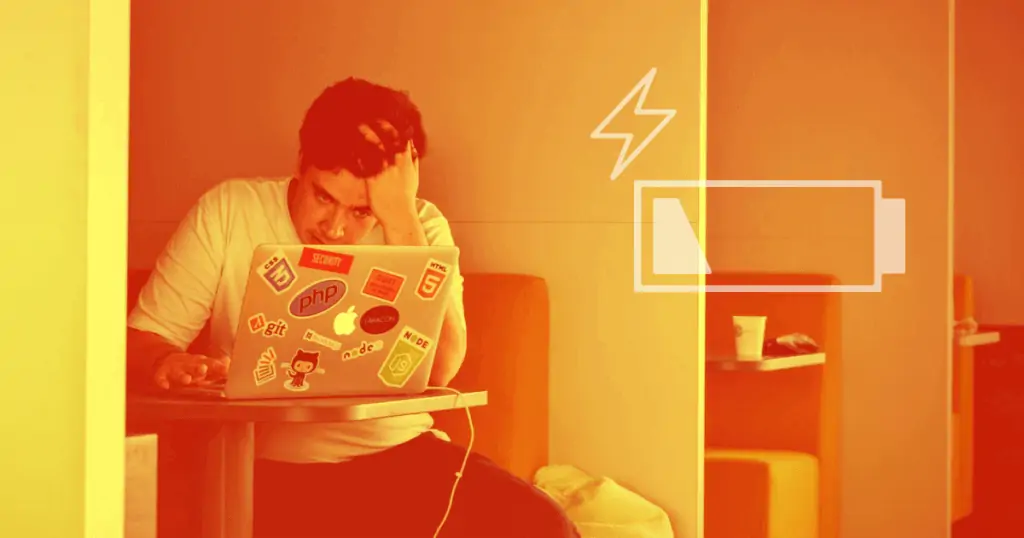 A man in a white T-shirt sits in front of a laptop computer covered in stickers. He has his head resting on his left hand, looking exhausted. There is an icon representing a low battery level to the right. Burnout is a serious problem for business owners.