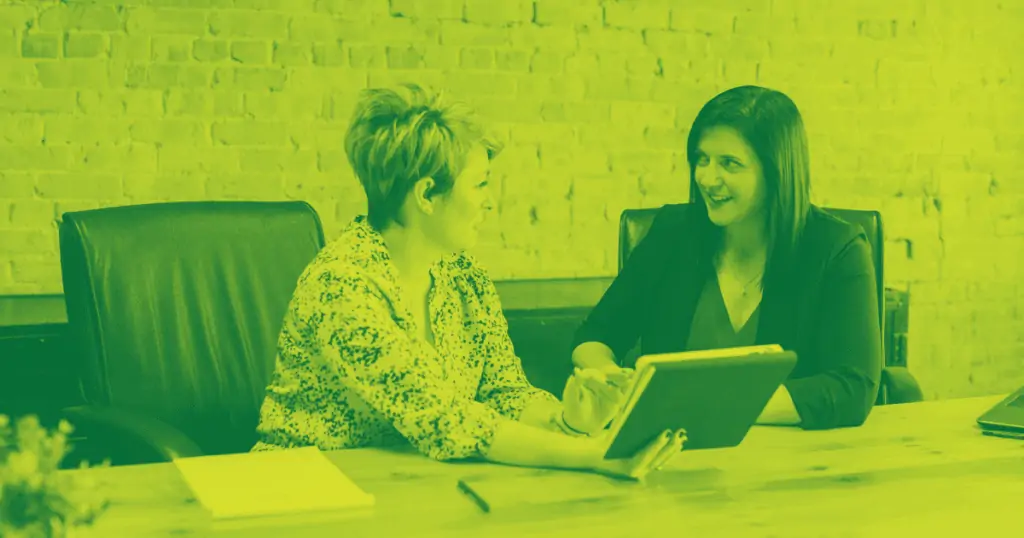 Two women are sitting at a table in front of a laptop computer, discussing a customer IEP. The photo appears in green and black.
