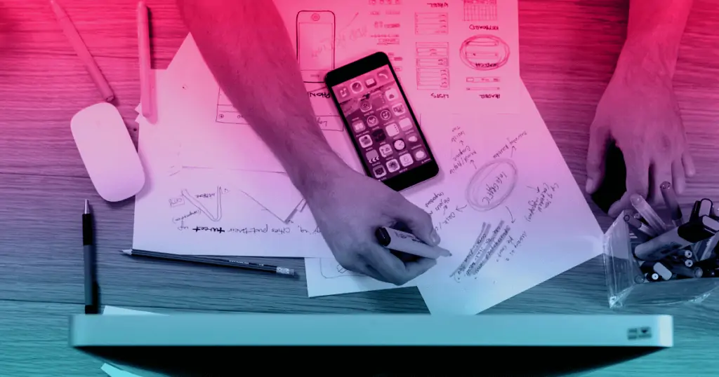 An overhead view of a person's desk. Their hands are visible, holding a highlighter. Next to a number of documents is a cell phone. The person may be working on an answer to the question, "What is digital marketing?"