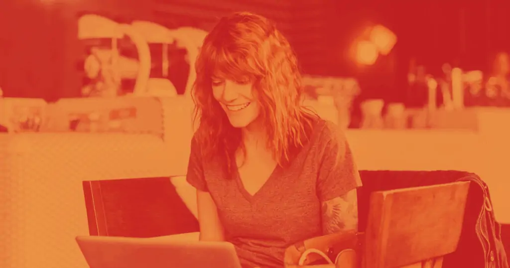 Pictured is a smiling woman working at a laptop computer at table surrounded by four chairs. She is in a cafe, working on content and branding for her business.