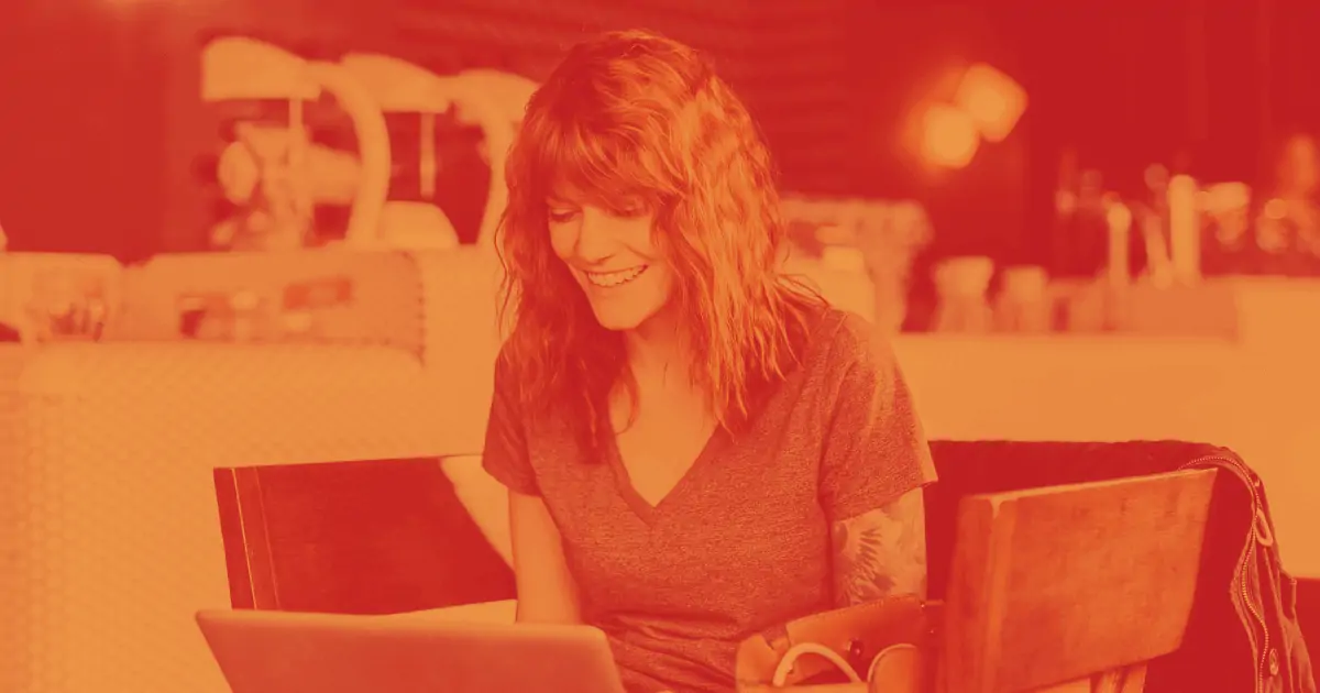 Pictured is a smiling woman working at a laptop computer at table surrounded by four chairs. She is in a cafe, working on content and branding for her business. 