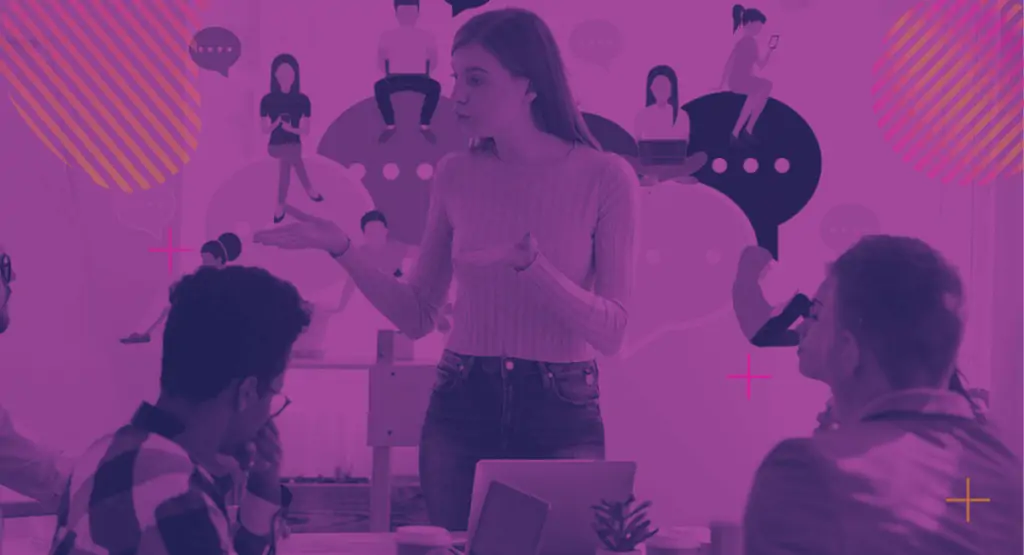 A young woman with long hair stands in front of a group of students, giving a presentation. The students are sitting at a table and have laptop computers open. The presentation could be centered around the benefits of customer-first branding and marketing.