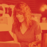 Pictured is a smiling woman working at a laptop computer at table surrounded by four chairs. She is in a cafe, working on content and branding for her business.