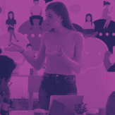 A young woman with long hair stands in front of a group of students, giving a presentation. The students are sitting at a table and have laptop computers open. The presentation could be centered around the benefits of customer-first branding and marketing.